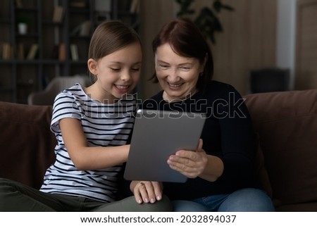 Joyful adorable little kid girl teaching smiling elderly mature grandmother using digital computer tablet software applications, sitting together on sofa at home, modern technology addiction concept.