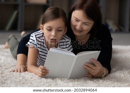 Affectionate middle aged elderly grandmother reading interesting paper book with joyful cute small kid granddaughter, enjoying relaxing carefree leisure weekend hobby activity together in living room.