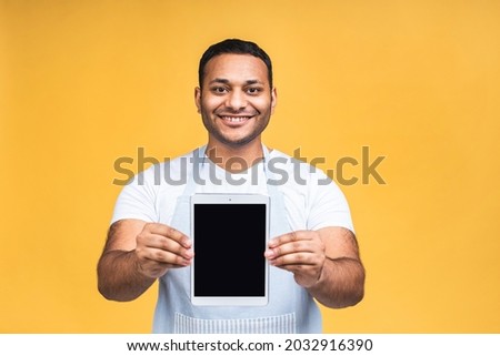 Studio picture of positive African American indian black chef or cook isolated over yellow background holding tablet and showing it blank screen with happy smile as if advising product or service.