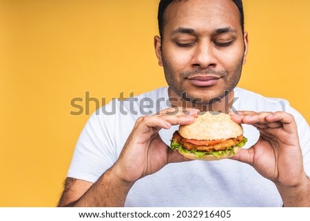 Young african american indian black man eating hamburger isolated over yellow background. Cook preparing burger.