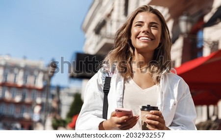 Happy blond woman, attractive girl holding takeaway cup, walking in city with smartphone in hand, looking aside. Girl going somewhere on street, drinking coffee and chat on mobile phone Royalty-Free Stock Photo #2032932557