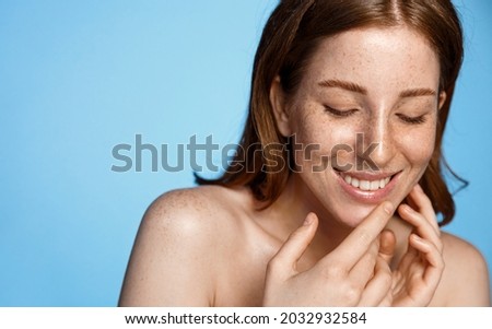 Skin care and facial treatment. Happy smiling girl with red hair, freckles and clean shiny skin, healthy face, touches her face and looks happy, blue background