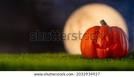 Halloween composition. An orange pumpkin with a creepy smile on green grass against a dark night sky and a huge white moon. Minimalism. Concept - Halloween celebration.