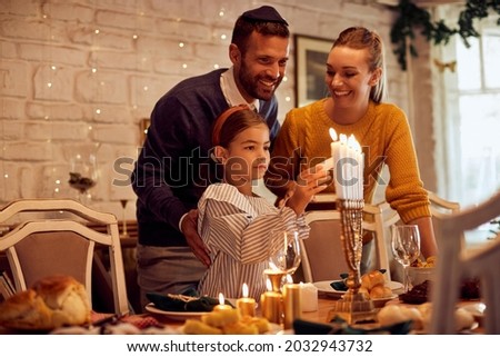 Happy parents with daughter lightning candles in menorah at dining table while celebrating Hanukkah at home. Royalty-Free Stock Photo #2032943732