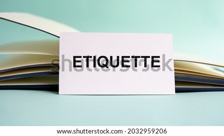 A white business card with ETIQUETTE text stands on a desk against the background of an open diary. Unfocused.