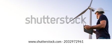Male engineer in the helmet controls and maintains the operation of a wind turbine using a laptop on the background of windmill. Banner of production of alternative and renewable energy concept. Royalty-Free Stock Photo #2032972961