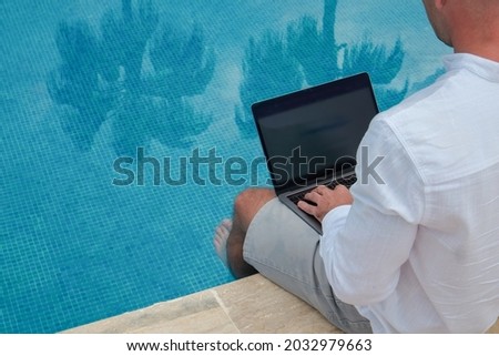 Young man wearing white linen shirt working on laptop by the pool, dipping his feet in a water. Male travel blogger typing on keyboard. Perks of being a freelancer. Close up, copy space, background.
