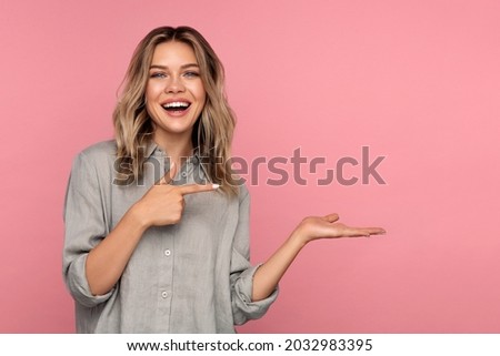 Excited young woman holding palm open for new product or sale presentation. Overjoyed happy blond girl with open mouth point finger at arm with copy space for promotion isolated over pink studio wall Royalty-Free Stock Photo #2032983395