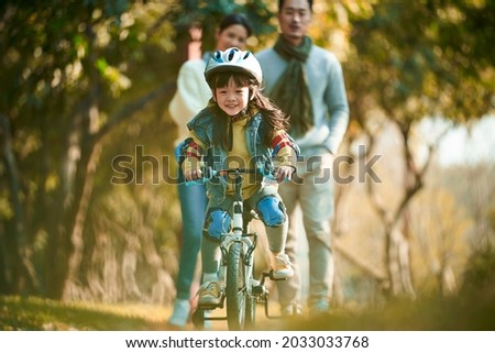 little asian girl with helmet and full protection gears riding bike in city park with parents watching from behind Royalty-Free Stock Photo #2033033768
