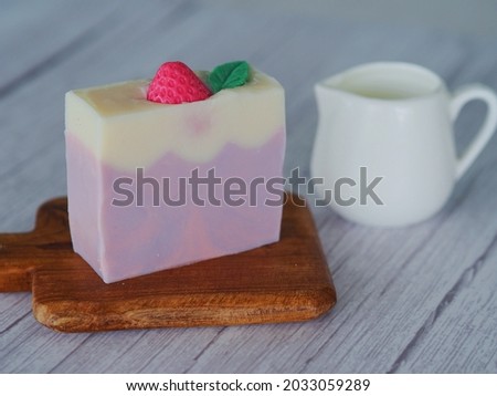 Side view handmade organic slices of purple pink cake shape with strawberry on top soap on white wooden table with milk cup. Selective focus. Melt and pour process. Cosmetic business concept.