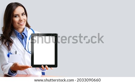 Portrait image of happy smiling female doctor showing tablet pc ipad touchpad with blank copy space area for slogan or text, over grey colour background. Medical center centre ad concept.