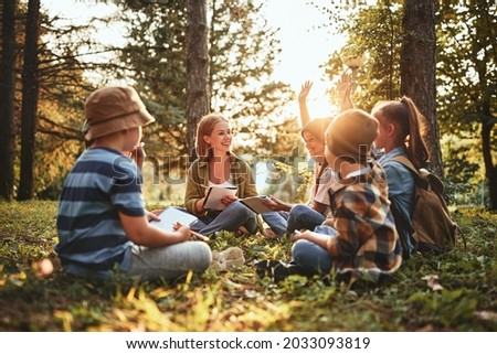 School children learning about ecology, nature and environment with teacher while sitting together on green grass in forest on sunny day, kids raising hands asking questions during outdoor lesson Royalty-Free Stock Photo #2033093819