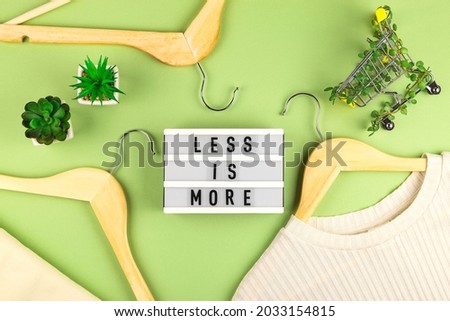 Less is more - written on lightbox next to Shopping cart entwined with plants on green background among wooden hangers. Conscious consumption slow fashion Zero waste concept. Top view flat lay