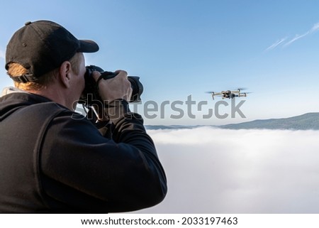 a male photographer takes pictures of a drone hovering in a thick fog in the air above the top of a mountain