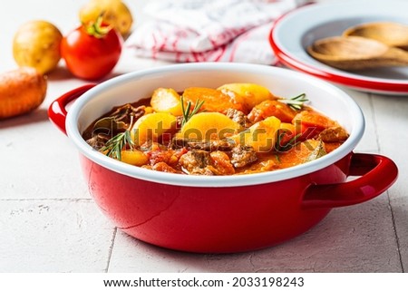 Beef stew with potatoes and carrots in tomato sauce in red pot, gray  background. Slow cooking concept. Royalty-Free Stock Photo #2033198243