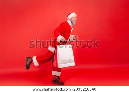 Full length side view portrait of elderly man with gray beard wearing santa claus costume moving running with shopping bags, presents for Christmas. Indoor studio shot isolated on red background.