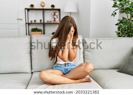Young brunette teenager sitting on the sofa at home with sad expression covering face with hands while crying. depression concept.  Royalty-Free Stock Photo #2033221382