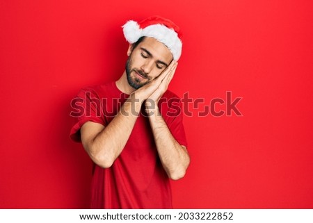 Young hispanic man wearing christmas hat sleeping tired dreaming and posing with hands together while smiling with closed eyes. 