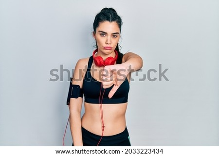 Young hispanic girl wearing gym clothes and using headphones looking unhappy and angry showing rejection and negative with thumbs down gesture. bad expression. 