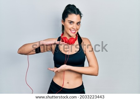Young hispanic girl wearing gym clothes and using headphones gesturing with hands showing big and large size sign, measure symbol. smiling looking at the camera. measuring concept. 