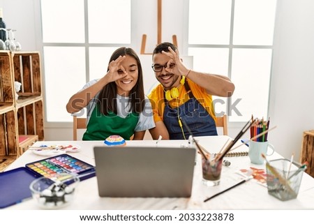 Young hispanic couple using laptop sitting on the table at art studio smiling happy doing ok sign with hand on eye looking through fingers 