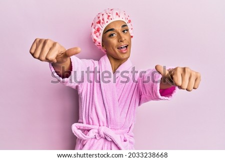 Hispanic man wearing make up wearing shower towel cap and bathrobe approving doing positive gesture with hand, thumbs up smiling and happy for success. winner gesture. 