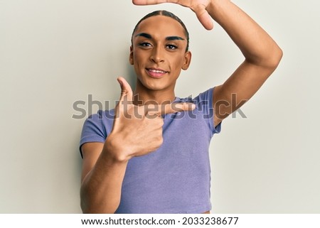Hispanic man wearing make up and long hair wearing casual t shirt smiling making frame with hands and fingers with happy face. creativity and photography concept. 