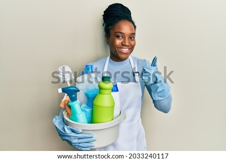 African american woman with braided hair wearing apron holding cleaning products smiling happy and positive, thumb up doing excellent and approval sign 