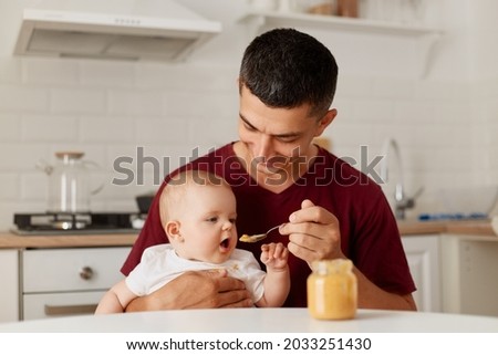Indoor shot of happy father sitting at table with little baby girl in arms and feeding daughter with fruit or vegetable puree, complementary feeding of child. Royalty-Free Stock Photo #2033251430