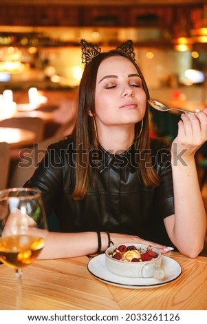 Portrait of young gorgeous woman drinking champagne in glass and looking with smile eating ice cream fruit dessert. Enjoying her leisure time inside a restaurant. Girl in a black dress with cat ears.