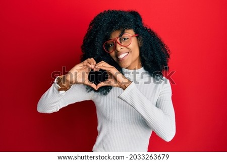 African american woman with afro hair wearing casual sweater and glasses smiling in love doing heart symbol shape with hands. romantic concept. 