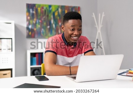 Trainee in office sits at desk in front of laptop monitor in background whiteboard, young dark skinned man talking on internet with clients, co-workers, uses webcam, video call, puzzled surprised face Royalty-Free Stock Photo #2033267264