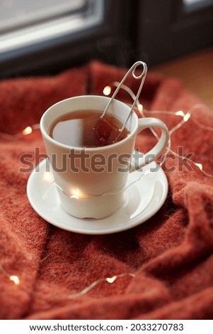 drinks and objects concept - cup with mesh tea infuser ball and garland lights on window sill at home