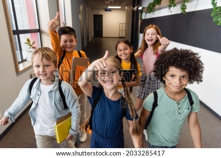 cheerful multicultural schoolchildren showing thumbs up in school corridor