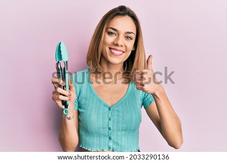 Young caucasian blonde woman holding silicone spatula smiling happy and positive, thumb up doing excellent and approval sign 