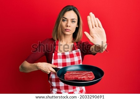 Young caucasian blonde woman wearing cook apron cooking meat on pan with open hand doing stop sign with serious and confident expression, defense gesture  Royalty-Free Stock Photo #2033290691