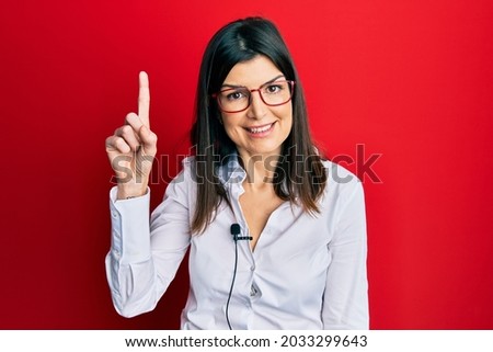 Young hispanic woman using lavalier microphone surprised with an idea or question pointing finger with happy face, number one 