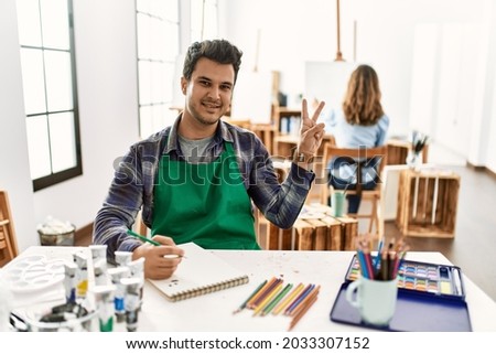 Young artist man at art studio smiling looking to the camera showing fingers doing victory sign. number two. 