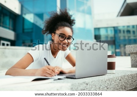 E-Learning. Cheerful African Student Girl Taking Notes At Laptop Learning Distantly Sitting Outdoor Near Urban College Building, Wearing Eyeglasses. Modern Online Education Royalty-Free Stock Photo #2033333864