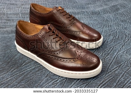 Formal golf wingtip sneakers on a blue textured leather background. The mens fashion footwear has a white outsole and is a fancy sneaker.