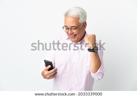 Middle age Brazilian man isolated on white background using mobile phone and doing victory gesture