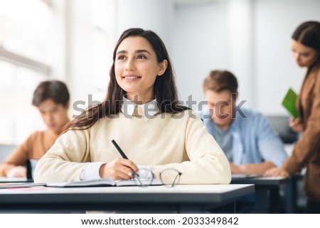 Portrait of smiling female university student holding pen writing in notebook doing examination or quiz test from teacher sitting at desk, attending classroom lesson, looking aside and thinking