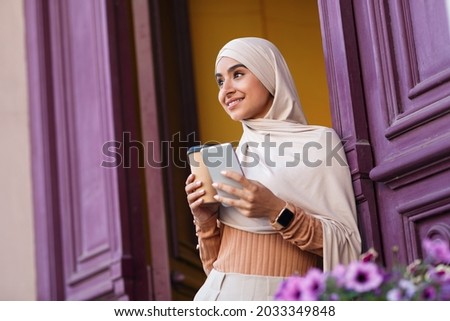 Good morning coffee on way to work, satisfied facial expression, enjoy outdoor. Smiling young muslim lady in hijab with smartphone and delicious takeaway coffee near front door of house in city