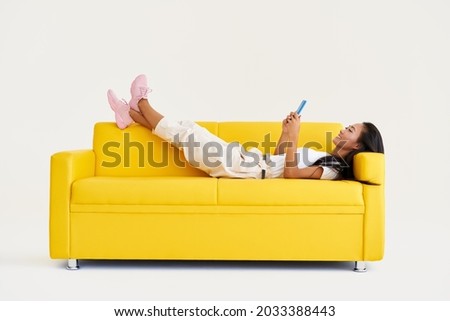 Smiling asian woman types text message on cell phone, enjoys online communication lying on yellow couch on white background. Technology, communication concept Royalty-Free Stock Photo #2033388443