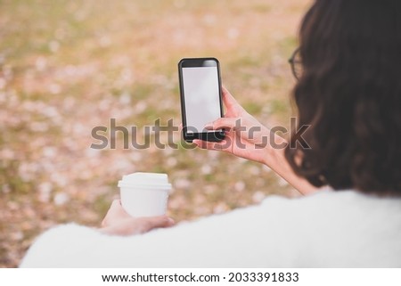 A young woman look and play on the mock up white blank screen vertical mobile smart phone in the public botanical garden park as hobby and recreation for relaxation, game and application concept