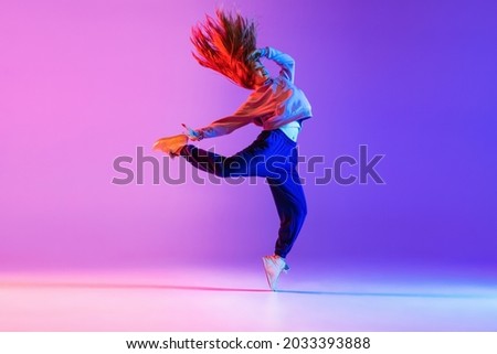 Dancing style. Young beautiful girl expressively making hip-hop tricks on gradient neon background. Youth culture, style and fashion. Concept of dance, youth, hobby, dynamics, movement, action, ad Royalty-Free Stock Photo #2033393888