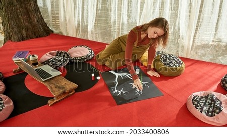 Young artist drawing a white tree on a5 paper while sitting on the red floor, surrounded with poufs, wearing brown overalls and burgundy blouse, drawing outside in a tent