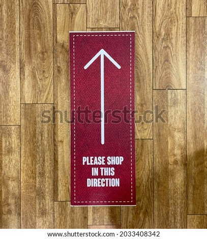 A retail shop sign with white lettering and arrow on burgundy background on a wood laminate floor says 'please shop in this direction'