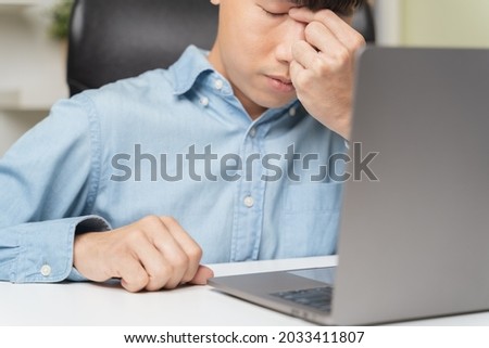 Stressed young Asian businessman, office worker massaging nose bridge feeling headache, eye strain, massaging to relieve pain from migraine, dizziness, blurry vision on overwork, using computer laptop Royalty-Free Stock Photo #2033411807