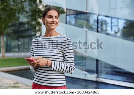 Photo of cheerful brunette woman spends free time at street holds mobile phone wears casual striped jumper sends messages blogs in social networks looks happily away poses outdoor blurred background